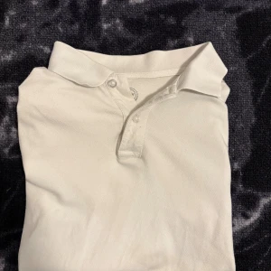 Vit pikétröja  - Klassisk vit pikétröja med krage och två knappar framtill. Tröjan är i bomull och har korta ärmar, perfekt för en clean och stilren look. Passar dig som gillar enkel och tidlös stil. Har antal fläckar