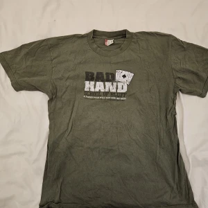 Olivgrön t-shirt från Levi's XL - Olivgrön t-shirt från Levi's i storlek XL känns  som L, med tryck på bröstet: 'BAD HAND SCHOOL OF BLUFF' och spelkortsgrafik. Klassisk passform, rund hals och korta ärmar. Skön bomullskvalitet, perfekt för dig som gillar poker eller coola statement-plagg.