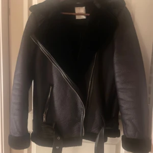 Svart teddyjacka från Pieces - Säljer en svart teddyjacka från Pieces med bred krage, dragkedja i silver och bälte i midjan. Jackan har mjukt foder, uppvikta ärmslut och två snedställda fickor med dragkedja. Perfekt för kalla dagar och riktigt snygg till jeans eller kjol.