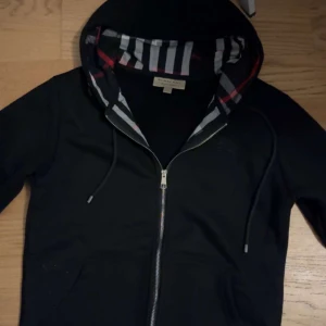 Svart Burberry hoodie med dragkedja - Svart hoodie från Burberry med klassiskt rutigt foder i huvan och snygga detaljer som silverfärgad dragkedja och snörning. Tillverkad i 100% bomull för skön känsla. Perfekt för dig som gillar stilrena och exklusiva plagg med ikonisk design.