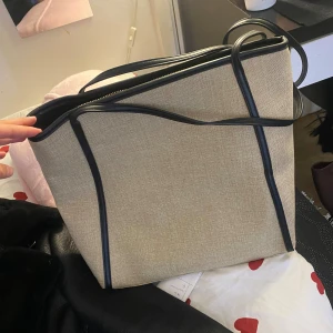 Beige axelväska med svarta detaljer - Stilren beige axelväska med svarta kanter och handtag. Väskan har en fyrkantig form och stängs med dragkedja upptill. Tillverkad i ett tygmaterial med detaljer i syntet, perfekt för att bära allt du behöver till skolan eller stan. Väger väldigt lätt köpt från Kappahl inte använd i jätte bra skick. 