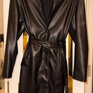 Svart skinnrock från Bershka XS - Snygg svart skinnrock från Bershka i storlek XS. Jackan har klassisk krage, knäppning med knappar och ett bälte i midjan. Tillverkad i syntetmaterial med slät finish och har praktiska fickor på sidorna. Perfekt för dig som vill ha en stilren och edgy look.