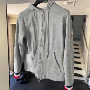 Grå Moncler hoodie med dragkedja - Snygg grå hoodie från Moncler med huva och snörning. Tröjan har hel dragkedja framtill, fickor och Moncler-logga på ärmen. Muddarna har coola ränder i rött, vitt och blått för extra stilpoäng. Perfekt för chill dagar och streetwear. Lite sönder syns på dragkedjan.
