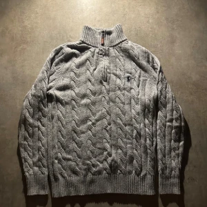 Grå stickad half zip tröja Polo Ralph Lauren - Säljer en grå stickad tröja från Polo Ralph Lauren med half zip och kabelstickat mönster. Tröjan har ribbad krage, muddar och nederkant samt långärmad design. Diskret broderad logga på bröstet. Perfekt för kyliga dagar.