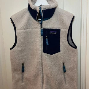 Beige fleeceväst från Patagonia S - Beige fleeceväst från Patagonia med marinblå detaljer och dragkedjor. Säljer då den inte kommer till användning. Hör av för frågor och funderingar