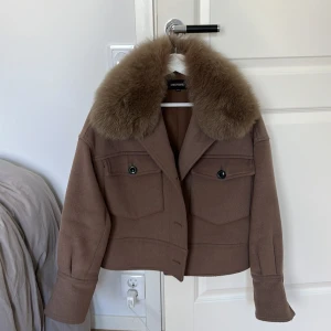 Brun jacka med päls från Meotine - Säljer en brun jacka från Meotine med stor fluffig pälskrage. Jackan har två stora fickor framtill med knappar och en croppad passform.