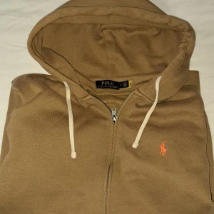 Brun hoodie från Polo Ralph Lauren - Snygg beige hoodie från Polo Ralph Lauren med dragkedja och huva. Tröjan har vita snören, två fickor framtill och den klassiska broderade loggan i orange på bröstet. Perfekt för chill dagar och enkel att matcha med jeans eller joggers.