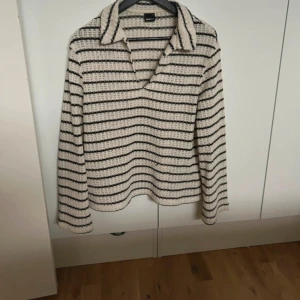 Randig stickad tröja med krage - Säljer en beige och svart randig stickad tröja med v-ringning och bred krage. Tröjan har lång ärm och en avslappnad passform, perfekt för lager på lager. Den grova stickningen ger en cool och trendig look.