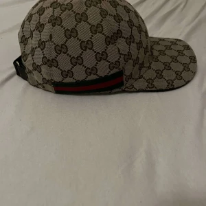 Gucci keps med GG-mönster ⚠️Helt Äkta! - Snygg beige keps från Gucci med klassiskt GG-monogram i brunt över hela kepsen. Har en grön och röd rand på sidan och justerbar rem baktill. Insidan är svart med Gucci-etikett. Perfekt för dig som gillar lyxiga accessoarer.