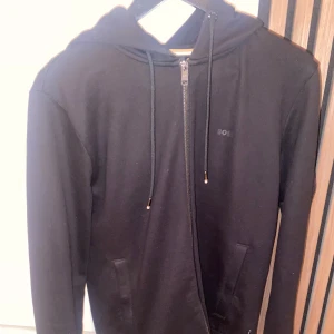 Svart hoodie från BOSS med dragkedja - Svart hoodie från BOSS med hel dragkedja framtill, snörning i huvan och två fickor. Stilren design med diskret logga på bröstet. Perfekt för en avslappnad och cool look.