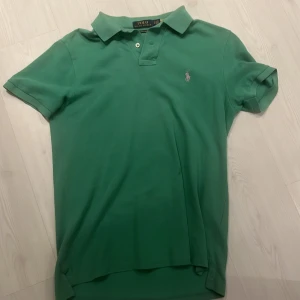 Grön pikétröja från Polo Ralph Lauren - Snygg grön pikétröja från Polo Ralph Lauren med klassisk krage och korta ärmar. Tröjan har den ikoniska broderade loggan på bröstet och är i bomull. Perfekt för en chill och stilren look.Har några fläckar men går att tvätta.
