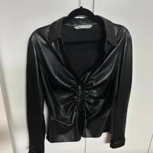 Svart skinnskjorta från Zara - Snygg svart skjorta i skinnimitation från Zara med lång ärm och cool snörning framtill. Skjortan har klassisk krage och en figurnära passform som ger en edgy vibe. Perfekt för dig som vill sticka ut med en trendig look.