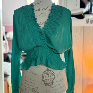 Grön blus med smock från SHEIN - Säljer en grön blus från SHEIN i storlek S. Blusen har smockdetaljer vid midjan och längs den djupa v-ringningen, samt volangkant nedtill. Långärmad och tillverkad i ett lätt, flowy material som ger en snygg siluett.