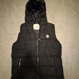 Moncler Väst - Svart dunväst från Moncler med huva och broderad logga på bröstet. Västen har quiltad design, två fickor med dragkedja och en snygg Moncler-text på huvan. Perfekt för lager-på-lager och streetstyle. Dragkedja framtill och coolt serietidningsmärke på insidan. Storlek är 13-14 år och passar 165-170-175 cm perfekt.