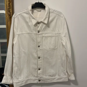 Vit jeansjacka med knappar - Säljer en stilren vit jeansjacka med klassisk krage och silverfärgade knappar framtill. Jackan har en bröstficka och raka sömmar för en clean look. Perfekt för dig som vill ha en enkel men snygg jacka till din outfit.