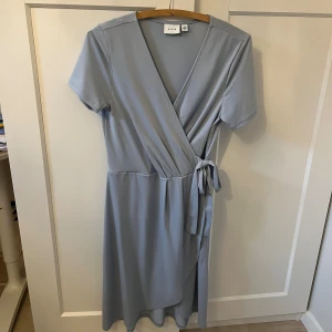Ljusblå omlottklänning från Vila - Superfin ljusblå omlottklänning från Vila med korta ärmar och knyt i midjan. Klänningen har v-ringning och en mjuk, flowy passform som ger en avslappnad men ändå stilren look. Använd ett fåtal gånger.