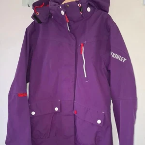 Lila vindjacka från McKinley - Snygg lila vindjacka från McKinley med huva och dragkedja. Jackan har stora fickor framtill. Perfekt för dig som vill sticka ut med färg.