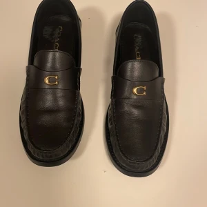 Svarta loafers från Coach i skinn - Snygga svarta loafers från Coach i mjukt skinn med rund tå och platt sula. På ovansidan sitter en guldfärgad metall-detalj med ett C, vilket ger en lyxig känsla. Klassisk och stilren design som passar till många outfits. Skorna köptes förra månaden från Boozt för 2050 kr.