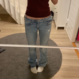 Ljusblå lågmidjade jeans med slitningar - Säljer ett par ljusblå jeans med låg midja och raka ben. Jeansen har slitna detaljer på knäna och klassiska fem fickor, varav bakfickorna har lock och knapp. Materialet är jeans och passformen är normal med lite bootcut-vibbar.