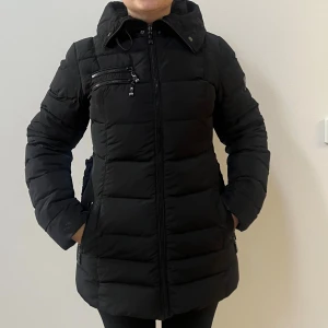 Svart pufferjacka från Hollies, stl 36 - Snygg svart pufferjacka från Hollies med hög krage, huva och dragkedjor både framtill och på bröstet. Jackan har markerad midja med resår baktill och flera praktiska fickor. Perfekt för kalla dagar och riktigt skön att bära.