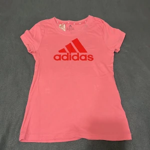 Rosa Adidas t-shirt med rött tryck - Snygg rosa t-shirt från Adidas med klassiskt rött logotryck på bröstet. Modellen har rund hals och korta ärmar, perfekt för dig som gillar sportig och enkel stil. Materialet är mjuk bomull som känns skönt mot huden.