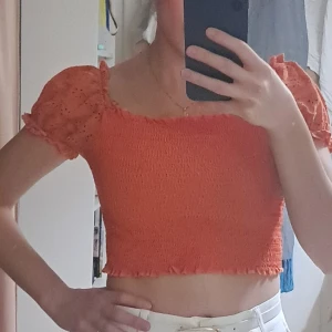 Orange croppad blus med puffärm - Superfin orange croppad blus med smockad kropp och korta puffärmar i broderad bomull. Söt volangkant upptill och nertill, samt elastisk passform som sitter snyggt. Perfekt för dig som gillar färg och trendiga detaljer.