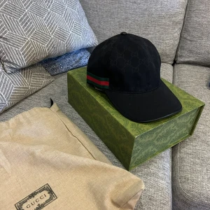 Svart keps från Gucci med logomönster - Säljer en svart keps från Gucci med diskret GG-logomönster över hela kepsen. Klassisk böjd skärm och justerbar rem bak. Sidan har den ikoniska gröna och röda randen. Tillverkad i bomull och polyester, perfekt för dig som vill ha en stilren accessoar.