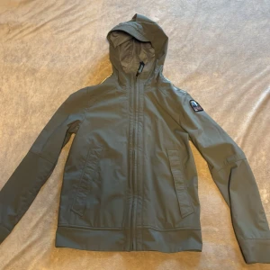 Grön softshelljacka från Parajumpers - Säljer en grön softshelljacka från Parajumpers med huva och dragkedja framtill. Jackan har två snedställda fickor, lång ärm och en snygg patch på ena axeln. Perfekt för dig som vill ha en stilren och funktionell jacka till vardags.