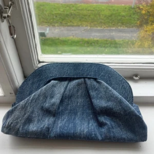 Blå denim clutch väska - Snygg kuvertväska i blå denim med veckad framsida och magnetlås. Väskan har en klassisk form och är fodrad med svart tyg invändigt. Perfekt för att bära det viktigaste och ge din outfit en cool touch.