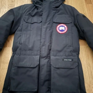 Svart parkajacka från Canada Goose - Säljer en svart parkajacka från Canada Goose med huva och stora fickor framtill. Jackan har klassisk logotyp på bröstet och praktiska detaljer som ficklock och dragkedja. Perfekt för kalla dagar och har en stilren look.