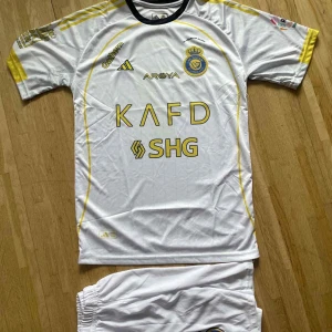 Al Nassr Ronaldo 7 fotbollströja Adidas - Vit Al Nassr fotbollströja med gula detaljer, tryckta sponsorer och klubbmärke. Nummer 7 och Ronaldo på ryggen. Klassisk Adidas-design med korta ärmar och rund hals. Tillverkad i lätt och ventilerande material, perfekt för match eller träning.