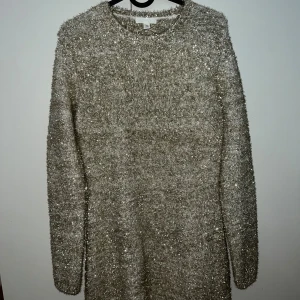 Glittrig stickad klänning från H&M - Superfin glittrig stickad klänning från H&M i beige och silver. Klänningen har lång ärm, kort längd och en snygg öppen rygg. Perfekt för dig som vill sticka ut med en mysig men ändå glam look. Materialet är mjukt och skimrande med glittertrådar.