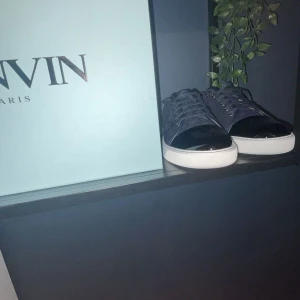 Lanvin sneakers i mörkblå mocka - Snygga sneakers från Lanvin i mörkblå mocka med svart lackad tå och vit platt sula. Skorna har snörning och en rund tå, vilket ger en clean och lyxig vibe. Perfekta för dig som gillar exklusiva detaljer och stilren design. Allt OG följer med 