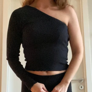 Svart oneshoulder topp Gina Tricot - Säljer en svart ribbad oneshoulder topp från Gina Tricot. Toppen har en lång ärm och croppad passform med vågig kant nedtill. XS stretch
