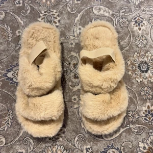 Beige fluffiga sandaler strl 39 - Supermysiga sandaler i beige med fluffigt pälsmaterial och två breda remmar över foten. De har en platt sula i svart gummi och en elastisk rem för extra komfort. Perfekta för chill dagar hemma eller när du vill ha något riktigt mjukt på fötterna.