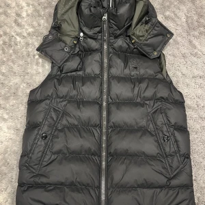 Svart dunväst från G-Star RAW - Stilren svart dunväst från G-Star RAW med huva, dragkedja framtill och två snedställda fickor med knapp. Västen har quiltad design och diskret logotyp på bröstet. Perfekt för lager-på-lager och snygg till streetwear. NYPRIS 2099 KR 