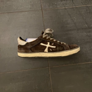 Premiata bruna sneakers i mocka - Säljer ett par bruna sneakers från Premiata med vit logga på sidan och detaljer i beige. Skorna har snörning och är tillverkade i mjuk mocka med en platt sula. Insidan och hälen har en ljusare, krackelerad look som ger extra karaktär. Perfekt för dig som gillar streetstyle. Sulan lagad nyligen!