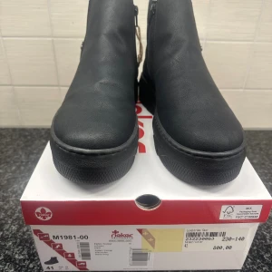 Svarta boots från Rieker, storlek 41 - Säljer ett par svarta boots från Rieker med rund tå och platt sula. Skorna har dragkedja på sidan och elastisk panel för enkel påtagning. Lätta och bekväma med fleecefoder inuti och diskret logga på sidan. Perfekta för höst och vinter. Helt nya i sin förpackning 