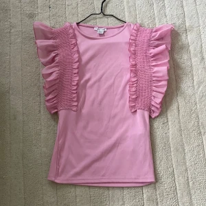 Rosa volangblus från Amisu XS - Supersöt rosa blus från Amisu i storlek XS. Toppen har breda volangärmar med smockdetaljer och är tillverkad i ett mjukt syntetmaterial. Säljs då den inte kommer till användning😊💗