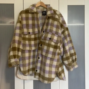 Rutig teddy-overshirt från Zara - Mysig oversized overshirt från Zara i teddy-material med rutigt mönster i beige, lila och brunt. Skjortan har stora bröstfickor med knappar, klassisk krage och långa ärmar med knapp vid ärmslut. Perfekt för lager-på-lager och chill stil.