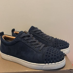 Mörkblå Christian Louboutin sneakers - Säljer ett par mörkblå sneakers från Christian Louboutin med coola nitar på tån och klassisk röd sula. Skorna har snörning, rund tå och är tillverkade i mocka med vit platt sula. Perfekt för dig som vill sticka ut med exklusiv design och edgy detaljer.