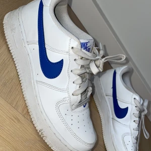 Nike Air Force 1 vit/blå sneakers stlk 37,5 - Nike Air Force 1 sneakers i vitt skinn med blå swoosh och detaljer. Klassisk låg modell med snörning och perforerad tå för extra ventilation. Platt sula med tydligt mönster och rund tå. Perfekt för dig som gillar stilrena och sportiga sneakers.