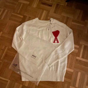 Vit stickad tröja från Ami Paris - Vit stickad tröja från Ami Paris med röd hjärtlogga och bokstaven A på bröstet. Tröjan har rund halsringning, ribbade muddar och är långärmad. Perfekt för dig som vill ha en clean look med en snygg detalj framtill.