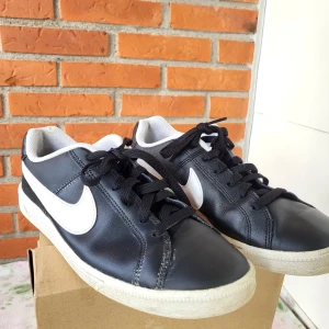 Svarta Nike sneakers strl 42 - Snygga svarta Nike sneakers med vit swoosh och vit yttersula. Klassisk låg modell med rund tå och snörning. Ovandel i skinn och insida i mesh för skön komfort. Perfekt för dig som gillar stilrena och sportiga skor.