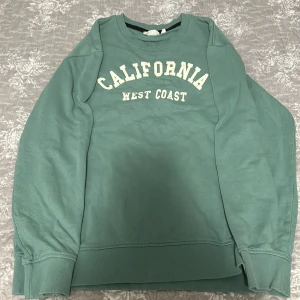 Grön sweatshirt California Kappahl - Cool grön sweatshirt från Kappahl med trycket 'California West Coast' i vitt framtill. Tröjan har rund hals, långa ärmar och breda muddar vid ärmslut och nederkant. Perfekt för en avslappnad och trendig look.