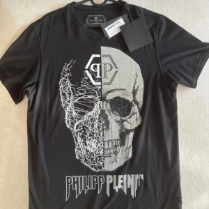 Svart t-shirt med döskalle från Philipp Plein - Cool svart t-shirt från Philipp Plein med stor döskalle framtill, där ena halvan är glittrig med paljetter och den andra har ett grafiskt mönster. T-shirten har korta ärmar och rund hals. Loggan är tryckt under motivet. Perfekt för dig som gillar edgy streetstyle. Aldrig använd, lappar kvar. Köpt i Marbella