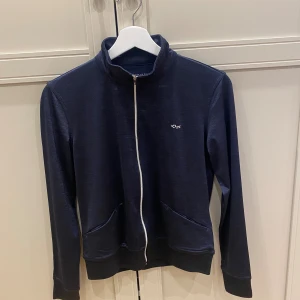 Träningströja från Röhnisch - Mörkblå ziptröja från Röhnisch med vit dragkedja och broderad logga på bröstet. Snygg brodyr i vitt på ryggen. Tröjan har hög krage, två fickor fram och ribbade muddar. 