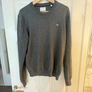 Mörkgrå GANT tröja i bomull och ull - Snygg mörkgrå tröja från GANT med broderad logga på bröstet. Klassisk rund halsringning och ribbade muddar. Tillverkad i en mjuk mix av bomull och ull som gör den både bekväm och stilren. Perfekt till jeans eller chinos. Mycket bra skick, har bara använts några gånger 