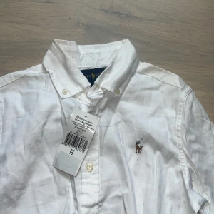 Vit skjorta från Ralph Lauren - Vit skjorta från Ralph Lauren med klassisk button-down krage och broderad logga på bröstet. Skjortan har lång ärm, knappar framtill och är gjord i mjuk bomull. Perfekt för dig som gillar stilrena och tidlösa plagg med snygga detaljer. Orginal. Aldrig använd. 
