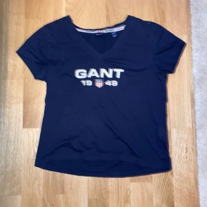 Mörkblå GANT t-shirt med v-ringning - Mörkblå t-shirt från GANT med v-ringad hals och tryck framtill. T-shirten är kortärmad och gjord i bomull. Tröjan har en kort passform. Inga hål eller andra defekter